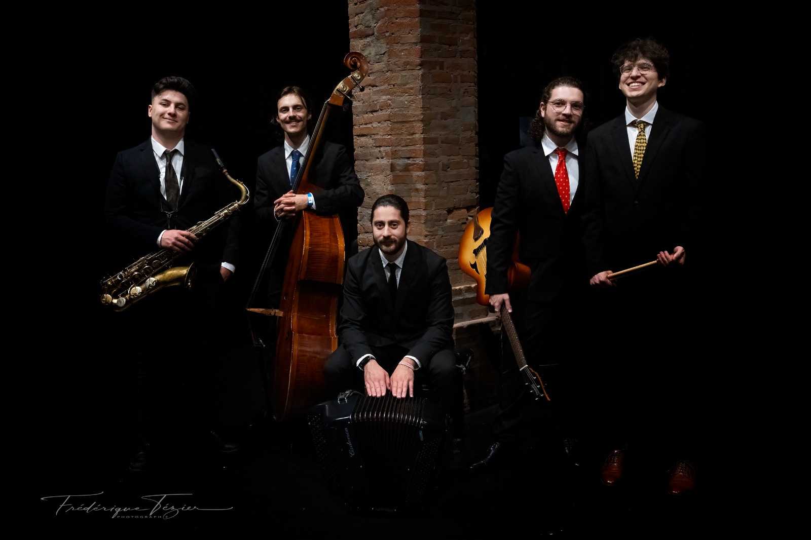 Un groupe de cinq musiciens masculins, un quintet de jazz, posant pour une photo. Ils sont habillés de manière formelle en costumes, tenant divers instruments, notamment un saxophone, une contrebasse, un accordéon et une guitare, avec un membre tenant des baguettes de batterie. L'arrière-plan est sombre avec un pilier en briques visible.